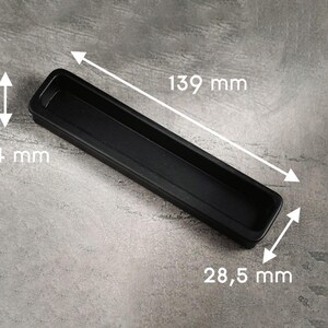 Può includere: Una maniglia per mobili rettangolare nera con bordi arrotondati, misura 139 mm di lunghezza, 28,5 mm di larghezza e 14 mm di profondità. La maniglia è posta su uno sfondo grigio, mostrando il suo design semplice e moderno.