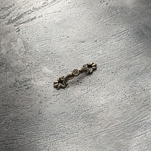 May include: A small, ornate, antique-style, bronze-colored drawer pull with a floral design.