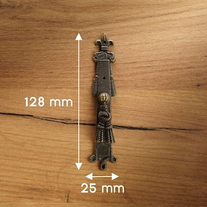 May include: An antique bronze-coloured cabinet pull, 128 mm long and 25 mm wide, with a fleur-de-lis design at the top and decorative rope and tassel elements. The hardware is set against a wooden surface.