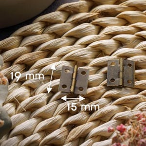 May include: Two small, antique brass-coloured hinges, each measuring 19 mm in height and 15 mm in width, are displayed against a woven, beige background. The hinges are open, showcasing their design and construction.