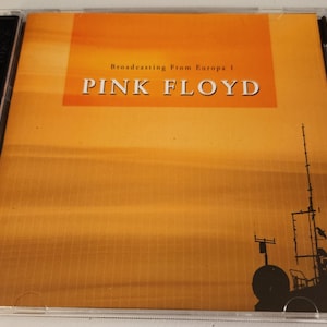 May include: A CD case with a yellow and orange background. The text "Broadcasting From Europa 1" and "PINK FLOYD" is printed on the front of the case. A silhouette of a ship with a tall antenna is in the background.