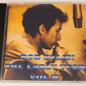 May include: A CD case with a yellow and orange background featuring a black and white photo of Bob Dylan in a recording studio. The text "Bob Dylan The Early Years Vol II" is printed in blue and white letters.