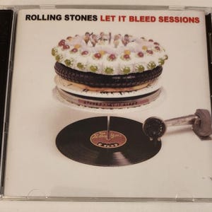 Könnte beinhalten: Ein CD-Gehäuse mit weißem Hintergrund und dem Text "Rolling Stones Let It Bleed Sessions". Das Bild zeigt einen Kuchen mit weißem Frosting und roten und grünen Dekorationen auf einem Plattenspieler.