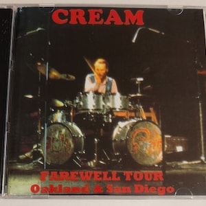 May include: A red and white CD case with a black border. The front of the case features a black and white photo of a drummer performing on stage. The text "CREAM" is in red at the top of the case. The text "FAREWELL TOUR Oakland & San Diego" is in red at the bottom of the case.