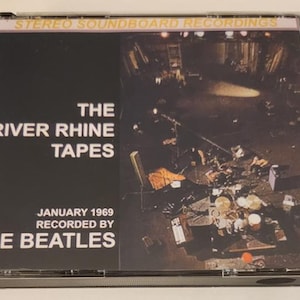 May include: A CD case featuring "The River Rhine Tapes" by The Beatles, recorded in January 1969. The cover has a black background with white text and a photograph of a stage. The top of the case reads "STEREO SOUNDBOARD RECORDINGS."