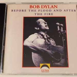 Puede incluir: Una portada de CD con una foto en blanco y negro de Bob Dylan cantando en un micrófono mientras sostiene una guitarra. El texto en la portada dice "Bob Dylan Before The Flood And After The Fire" con el logotipo de Luna Records en la parte inferior.