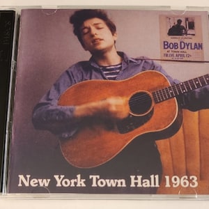 May include: CD case featuring Bob Dylan playing guitar. The album cover has a sepia-toned image of Dylan and the text "New York Town Hall 1963". A poster in the background advertises a concert.