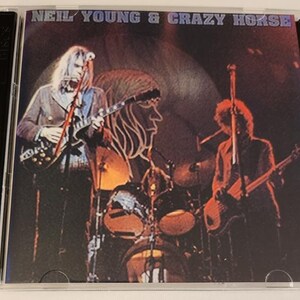 Könnte beinhalten: Ein CD-Gehäuse mit schwarzem Rücken und einem bunten Bild von Neil Young und Crazy Horse, die auf der Bühne auftreten. Der Text "Neil Young & Crazy Horse" ist oben auf dem Gehäuse in Weiß auf schwarzem Hintergrund gedruckt.