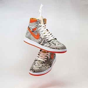 Op de afbeelding: Twee hoge sneakers met een slangenprint, oranje accenten en witte veters hangen in de lucht. De sneakers hebben een witte zool met een oranje rand en een groot oranje swoosh-logo aan de zijkant.