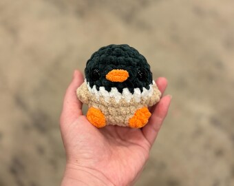 May include: A handmade crochet penguin with a dark green head, white chest, and tan body. The penguin has an orange beak and small black eyes. The penguin is being held in a hand.