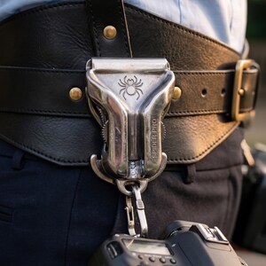 May include: A silver metal camera holster with a spider logo and the text SPIDER PRO engraved on the front. The holster is attached to a wide black leather belt with brass-colored rivets and a buckle, holding a camera via a metal carabiner clip.