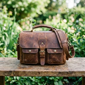 May include: A brown leather satchel with a top handle and adjustable shoulder strap. The bag features two front pockets with brass button closures and a buckle closure on the front flap. The bag is sitting on a wooden surface.