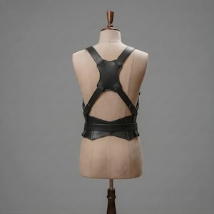 May include: A black leather harness featuring a central back panel with four straps extending over the shoulders and around the waist. The structure includes a horizontal waist belt connected to the back piece, creating a geometric, symmetrical design.
