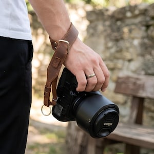 May include: A brown leather camera wrist strap with a metal buckle and a circular metal ring attachment. The strap is secured around a wrist and connected to a black Nikon camera body with a lens attached, featuring a black lens cap.