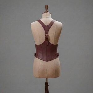 Puede incluir: Un cors&eacute; de cuero marr&oacute;n oscuro con un dise&ntilde;o de tiras en forma de Y en la espalda y una hebilla central de lat&oacute;n. La prenda est&aacute; confeccionada con costuras verticales e incluye correas laterales ajustables con herrajes de lat&oacute;n sobre un maniqu&iacute;.
