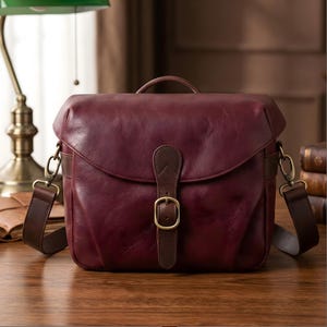 Peut inclure: Sac d'appareil photo en cuir bordeaux avec une poignée supérieure et une bandoulière réglable. Le sac est doté d'une fermeture à rabat avec une sangle en cuir marron et une boucle dorée. Le sac est posé sur une surface en bois.