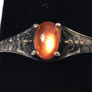 May include: A silver-toned ring featuring an oval-shaped, translucent orange cabochon stone set in a four-prong mount. The metal band displays a dark, oxidized finish with intricate, raised relief patterns extending from the central setting.