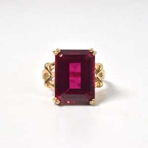 May include: A gold ring featuring a large, rectangular, emerald-cut deep red gemstone set in a prong setting. The band of the ring displays a decorative, openwork design with curved metal elements on both sides of the central stone.