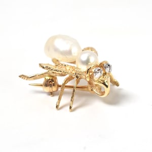 May include: A gold insect-shaped brooch featuring two white pearls forming the body and head. The insect has textured gold legs and wings, with two small clear stones set as eyes. A metal pin mechanism is visible on the underside of the brooch.
