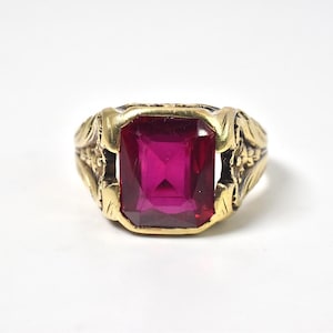 May include: A gold-toned ring featuring a large, rectangular, faceted deep pink gemstone set in the center. The metal band displays intricate, raised leaf-like carvings along the sides, providing a textured appearance to the shank of the ring.