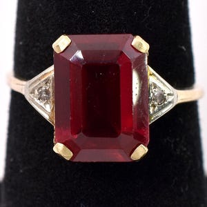 May include: A gold ring with a large, deep red emerald-cut gemstone and two smaller diamond accents.