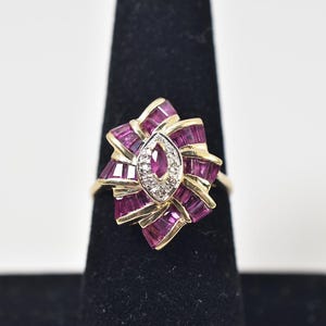May include: A gold ring featuring a central marquise-cut ruby surrounded by a halo of small round diamonds. The center stone is framed by a cluster of baguette-cut rubies arranged in a fan-like pattern, set in a yellow gold band structure.