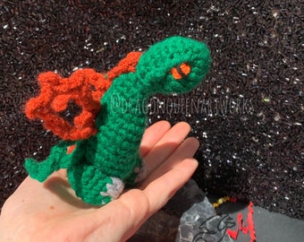 Puede incluir: Un dragón de crochet verde con alas naranjas y un ojo blanco. El dragón está sentado en una mano.