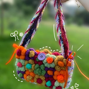 May include: A colorful, handmade crochet bag featuring a vibrant array of 3D flowers in shades of orange, purple, green, and yellow. The bag has a button closure and braided straps. Orange ribbons are tied on the sides.