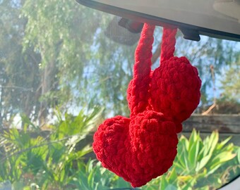 May include: Red crocheted heart-shaped car charm hanging from the rearview mirror of a car.