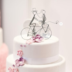 May include: A wire cake topper featuring two figures riding a bicycle, with the figure on the back wearing a veil and the front figure wearing a top hat. The rear wheel contains the text Olga and Max with a heart symbol between the names.