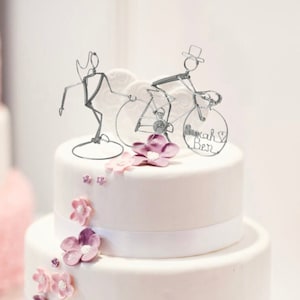 May include: A white two-tiered wedding cake decorated with purple flowers and a wire sculpture of a couple riding a bicycle. The bicycle's front wheel has the names "Sarah & Ben" written on it.