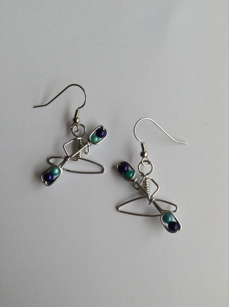 May include: A pair of silver wire earrings shaped like a person in a canoe with blue and teal beads.
