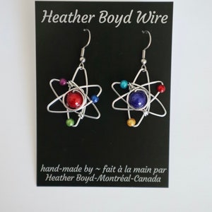 Puede incluir: Un par de pendientes de alambre plateado en forma de &aacute;tomo con una cuenta roja y una azul en el centro. Los pendientes est&aacute;n rodeados de cuentas m&aacute;s peque&ntilde;as de varios colores.
