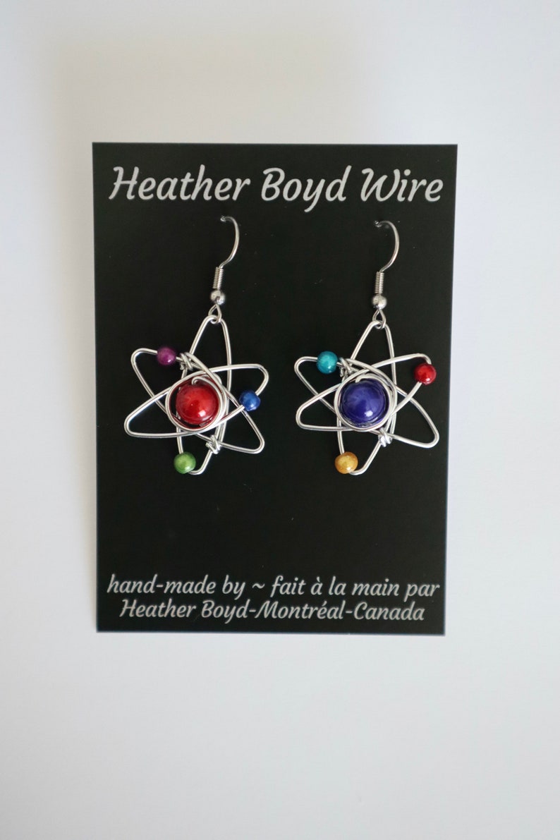 Puede incluir: Un par de pendientes de alambre plateado en forma de &aacute;tomo con una cuenta roja y una azul en el centro. Los pendientes est&aacute;n rodeados de cuentas m&aacute;s peque&ntilde;as de varios colores.