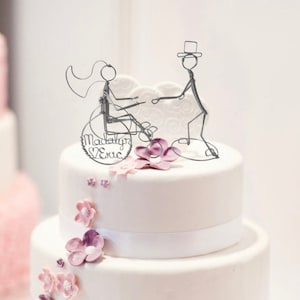 Pode incluir: Um bolo de casamento branco com uma escultura de arame de uma noiva e um noivo no topo. A noiva está em uma cadeira de rodas e o noivo está de pé ao lado dela. O bolo é decorado com flores rosa e roxas. O topo do bolo diz "Madelyn & Eric".