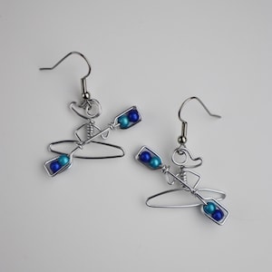 May include: A pair of silver wire earrings shaped like a person in a kayak with blue and turquoise beads for paddles.