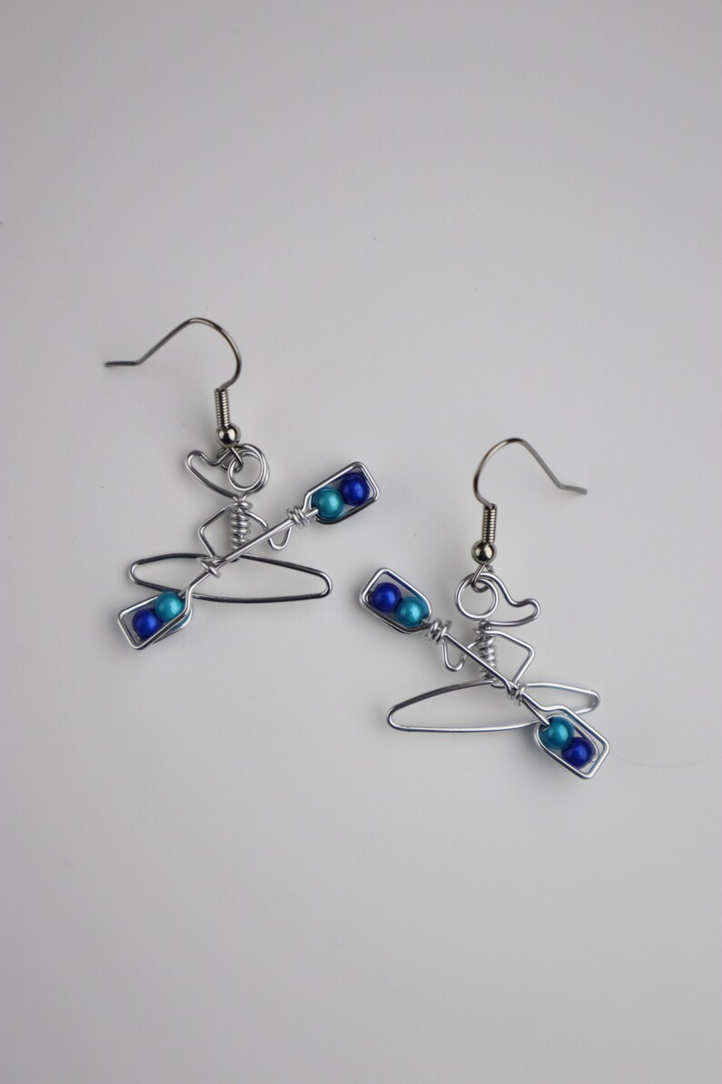 May include: A pair of silver wire earrings shaped like a person in a kayak with blue and turquoise beads for paddles.