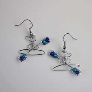 May include: A pair of silver wire earrings shaped like a person in a kayak with blue and turquoise beads for paddles.