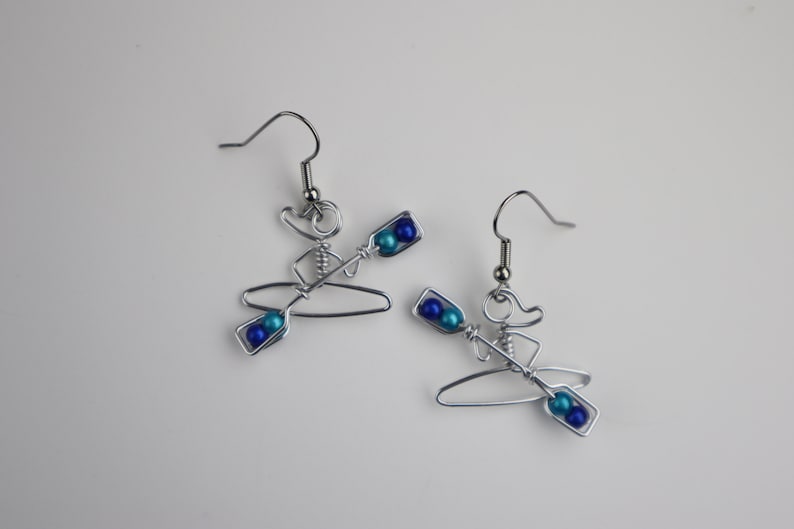 May include: A pair of silver wire earrings shaped like a person in a kayak with blue and turquoise beads for paddles.