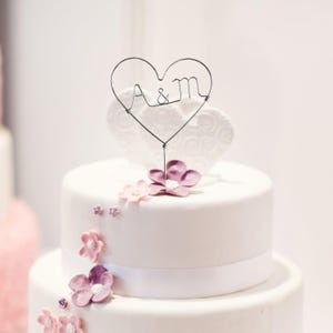May include: A white tiered wedding cake decorated with pink and purple flowers and a wire heart cake topper. The cake topper features the letters "A & M" inside the heart. A white ribbon encircles the cake.
