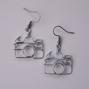 May include: A pair of silver wire camera earrings. The earrings are shaped like a camera with a lens and a shutter button. The earrings are hanging from silver hooks.