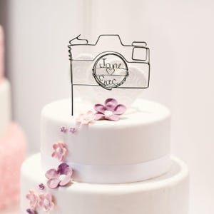May include: A white wedding cake decorated with pink and purple flowers. A wireframe camera cake topper with the names "Jane & Eric" is centered on the cake. The cake has a white ribbon around the base of the top tier.