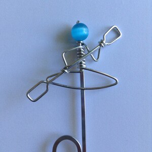 May include: A silver wire sculpture of a person in a kayak holding a paddle. The person's head is a blue bead.