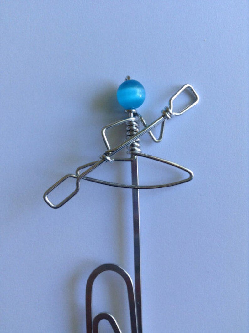 May include: A silver wire sculpture of a person in a kayak holding a paddle. The person's head is a blue bead.