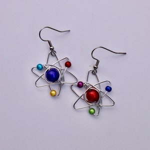 Puede incluir: Un par de pendientes de alambre plateado con forma de &aacute;tomos con cuentas de colores. Un pendiente tiene una cuenta azul en el centro y el otro tiene una cuenta roja en el centro.