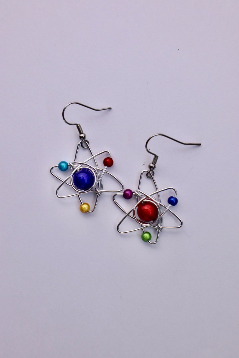 Puede incluir: Un par de pendientes de alambre plateado con forma de &aacute;tomos con cuentas de colores. Un pendiente tiene una cuenta azul en el centro y el otro tiene una cuenta roja en el centro.