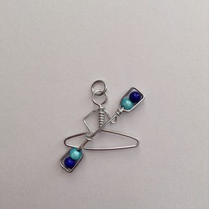 May include: A silver wire sculpture of a person in a kayak with blue and turquoise beads for paddles.