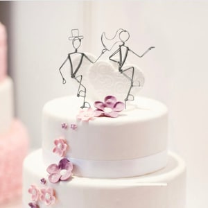 May include: A two-tiered white wedding cake decorated with purple and pink sugar flowers. A wire bride and groom topper sits on top of the cake.