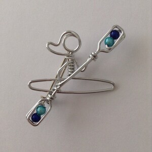 May include: A silver wire sculpture of a person in a kayak with blue and turquoise beads for the paddle.