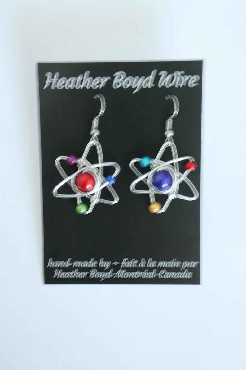 Puede incluir: Un par de pendientes de alambre plateado con forma de &aacute;tomos con cuentas de colores. Los pendientes est&aacute;n en una tarjeta negra con el texto "Heather Boyd Wire" y "hand-made by ~ fait &agrave; la main par Heather Boyd-Montr&eacute;al-Canada".